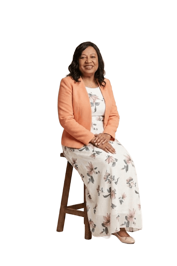 Nicky Menakaya, smiling, seated on a wooden stool in a floral dress and orange blazer